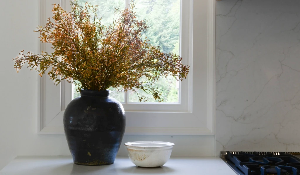 A vase of dried flowers sits on a counter next to a window in a home.